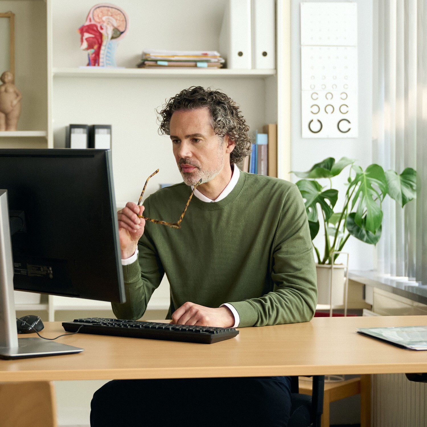 Huisarts kijkt geconcentreerd naar computerscherm terwijl hij zijn bril vasthoudt